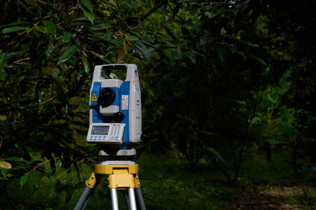 Terengganu, Malaysia - January 20, 2025 : A professional total station surveying instrument, mounted on its tripod, stands prominently in a lush, green outdoor environment. The bright sunlight illuminates the details of the precision equipment, including its control panel and various sensors. The natural, leafy background suggests a setting for land surveying, construction site preparation, or mapping projects in the field.の写真素材