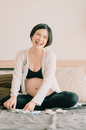 Young beautiful Asian pregnant woman lying in bed and choosing baby socks on bed - selective focus...