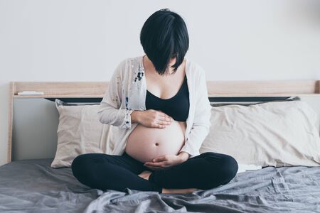 Young beautiful Asian pregnant woman lying in bed and touching her belly at home - selective focus point and vintage  styleの写真素材
