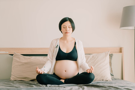 Young beautiful Asian pregnant woman meditating or doing yoga exercise at home. Relaxation yoga lotus pose. - selective focus point and vintage  styleの写真素材