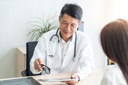 Asian senior doctor and patient are discussing something while sitting at the tableの写真素材