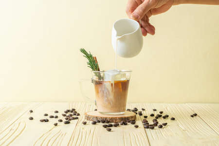 pouring milk in black coffee glass with ice cube, cinnamon and rosemary on wood backgroundの写真素材