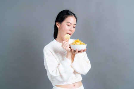 Young Asian woman in white sweater eatting potato chips on grey backgroundの写真素材