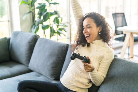 Laitin woman playing videogames with hands holding joystick on sofa at homeの写真素材