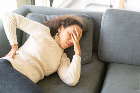 Latin woman lying down on sofa at home with backache feeling