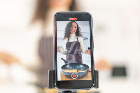 Latin woman shooting video and cooking at the kitchen for share on blogの写真素材