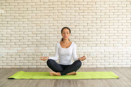 Latin woman practicing yoga on mat with white brick backgroundの写真素材