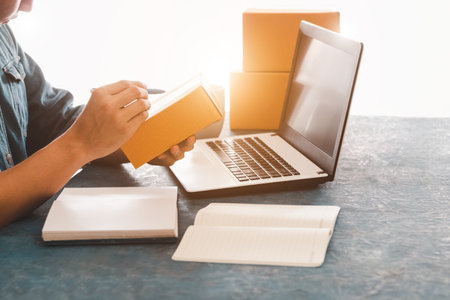 Young asian man Writing the customer's address on the brown storage box, business owners working in home packaging offices, E-commerce Online Shopping or Freelance Work Concept.の写真素材