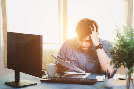 Young Asian men are stressed out in the business office. Looking at the expense bill and the other hand on the head, Thinking of making money to pay off all credit card debts and all bills. Concept of financial problems.の写真素材