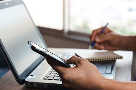 young man using  laptop computer and  mobile phone When looking for financial information in business, work at the desk. Writing with a pen, studying remotely from home and working from home.の写真素材