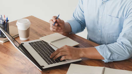 young man using laptop computer and mobile phone When looking for financial information in business, work at the desk. Writing with a pen, studying remotely from home and working from home.の写真素材