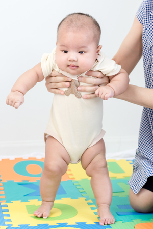 Portrait of a little adorable newborn infant baby girl standing up with mother support on colorful eva foam and looking in camera indoorsの写真素材