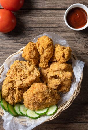 Crispy fried chicken in wicker basket serve with cucumber, tomato and ketchup on wooden background....
