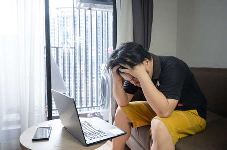 Stressed asian businessman sitting on a sofa at home and working on a laptop. tired man work from...