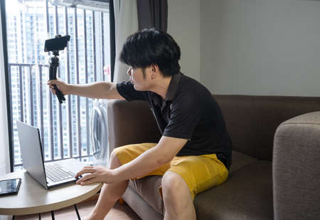 Asian man recording video for internet vlog while sitting on a sofa at home. Live broadcast and Blogger lifestyle concept.の写真素材