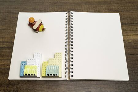 One red and yellow wooden airplane toy on blank white notebook with two ceramic buildingsの写真素材