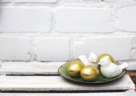 Golden Easter eggs and white porcelain birds in herbal green plate on white desk in front of brick...