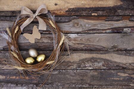 Rustic branch wreath with gold Easter eggs, feathers, butterfly and bow on dark brown wooden background. Copy space. Top view. Place for textの写真素材
