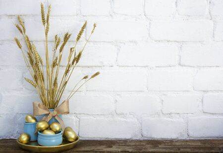 Easter decoration with gold eggs in blue pots with dry dyed wheat spikes on wooden table near white brick background. Horizontal, copy space. Place for textの写真素材