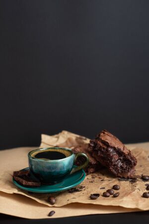 Dark green coffee cup and brownies with coffee beans and piece of chocolate on craft paper on black background. Healthy gluten free carob cake with cup of chicory. Vertical, copy spaceの写真素材