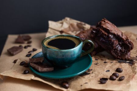 Dark green coffee cup and brownies with coffee beans and piece of chocolate on craft paper on black background. Healthy gluten free carob cake with cup of chicory. Vertical, copy spaceの写真素材
