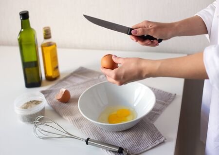 Female hands crack egg into bowl cooking omelet. Steel whisk, oil and vinegar bottles, salt and napkin on white table.の写真素材