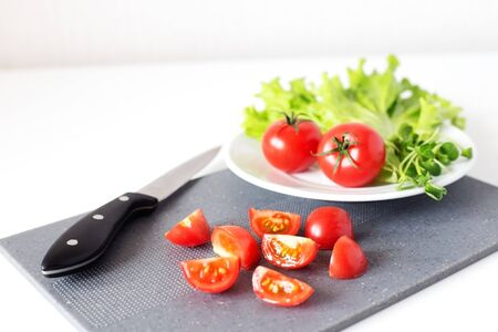 Slices of cherry tomatoes on grey chopping board with a knife on white background. Tomatoes with green salad on white plate. Close upの写真素材