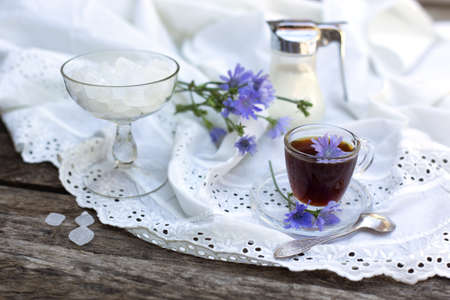 Healing drink. A transparent cup of chicory, jug of soy milk, sugar, with rustik napkin and blue flowers on a wooden background.の写真素材