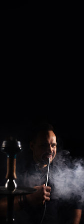 Vertical banner with a man smoking traditional hookah pipe. Man exhaling smoke on black background with copy space. Smoking concept for hookh bar. Place for textの写真素材
