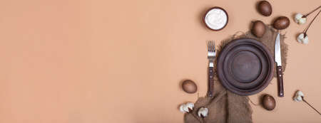 Horizontal banner with ceramic brown plate on sustainable cloth with Easter eggs, bowl with white cream sauce and cotton flowers on beige background with copy space. Zero waste card. Eco-friendlyの写真素材