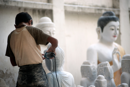 Burmese man use tool carving a large marble Buddha statue, outdoors. Man is surrounded with several other statues in Mandalay city,Central of Myanmar.のeditorial素材