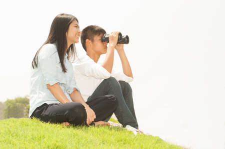 Asian young boy and girl sitting on grass using binoculars .の写真素材