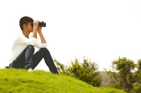 Asian young boy sitting on grass using binocularsの写真素材