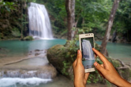 Hand of photographer with smart phone shooting image on blurred waterfall background.の写真素材