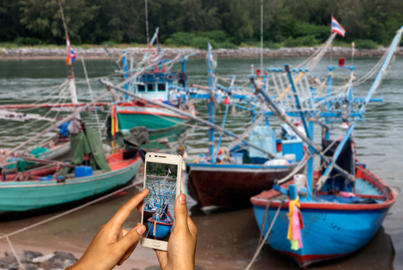 Hand of photographer with smart phone shooting image on blurred fishing boat on beach background.の写真素材