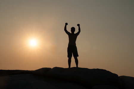Positive male holding hand up and expressing gladness while standing on stone in Sunset.の写真素材