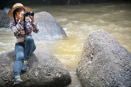 Young Asian traveler woman looking a binoculars in happiness and sitting on a rock of waterfall.の写真素材