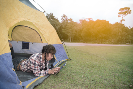 Beautiful smiling Asian woman lying down and looking tablet at the entrance of a blue - yellow camping tent.の写真素材