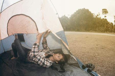 Beautiful smiling Asian woman lying down and looking tablet at the entrance of a blue - yellow camping tent.の写真素材