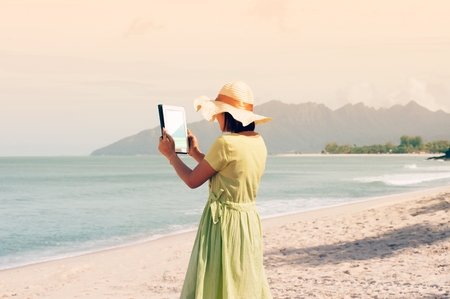 Young asian woman standing on the beautiful nice blue beach with white sand and taking photo with tablet.の写真素材