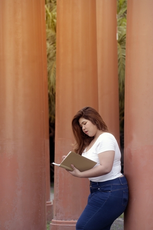 Portrait of Asian pretty smiley face fat woman pose and writing at a booklet on group of pole in the park.の写真素材