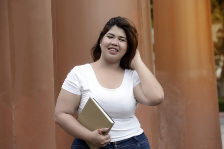 Portrait of Asian pretty smiley face fat woman pose standing and hold a booklet on group of pole in the park.の写真素材