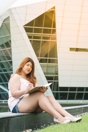 Portrait of Asian pretty smiley face fat woman pose ,holding booklet and writing near a pool front of building.の写真素材