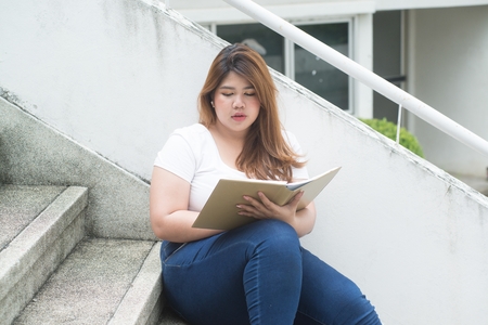 Portrait of Pretty Asian smiley fat woman sit and writing at staircase of building.の写真素材