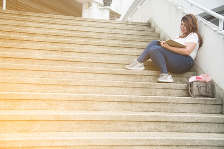 Portrait of Pretty Asian smiley fat woman sit and writing at staircase of building.の写真素材