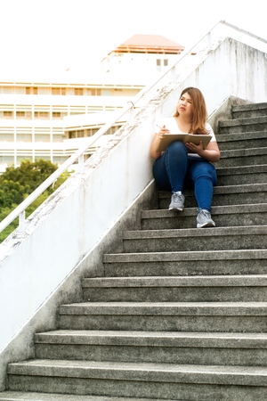 Portrait of Pretty Asian smiley fat woman sit and thinking at staircase of building.の写真素材