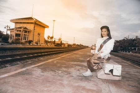 The lady in Middle Thai classical Thai traditional dress suit sitting and hold a paper fan is posing at railway station.の写真素材