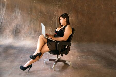 Asian plus size sexy , fat and overweight long hair model female in black dresses on brown studio background pose sitting use laptop.の写真素材