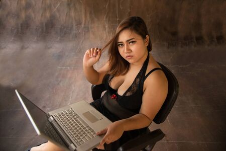 Asian plus size sexy , fat and overweight long hair model female in black dresses on brown studio background pose sitting use laptop.の写真素材