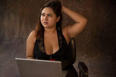 Asian plus size sexy , fat and overweight long hair model female in black dresses on brown studio background pose sitting use laptop.の写真素材